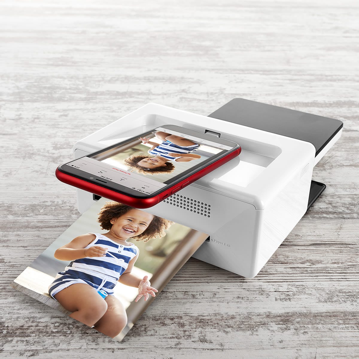 A compact photo printer with a smartphone dock prints a photo of a child in a striped romper. The phone, displayed in a red case, shows the same image on its screen, indicating a direct printing process from the device.