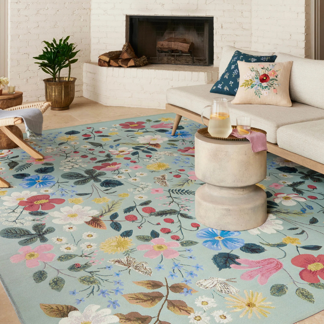 A 6' x 9' area rug features a colorful floral design with various blossoms and leaves on a light blue background, complementing a modern living room with a white couch, small tables, and a potted plant.