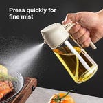 A hand is pressing the lid of a transparent oil dispenser, spraying a fine mist onto a pan of cooking food.