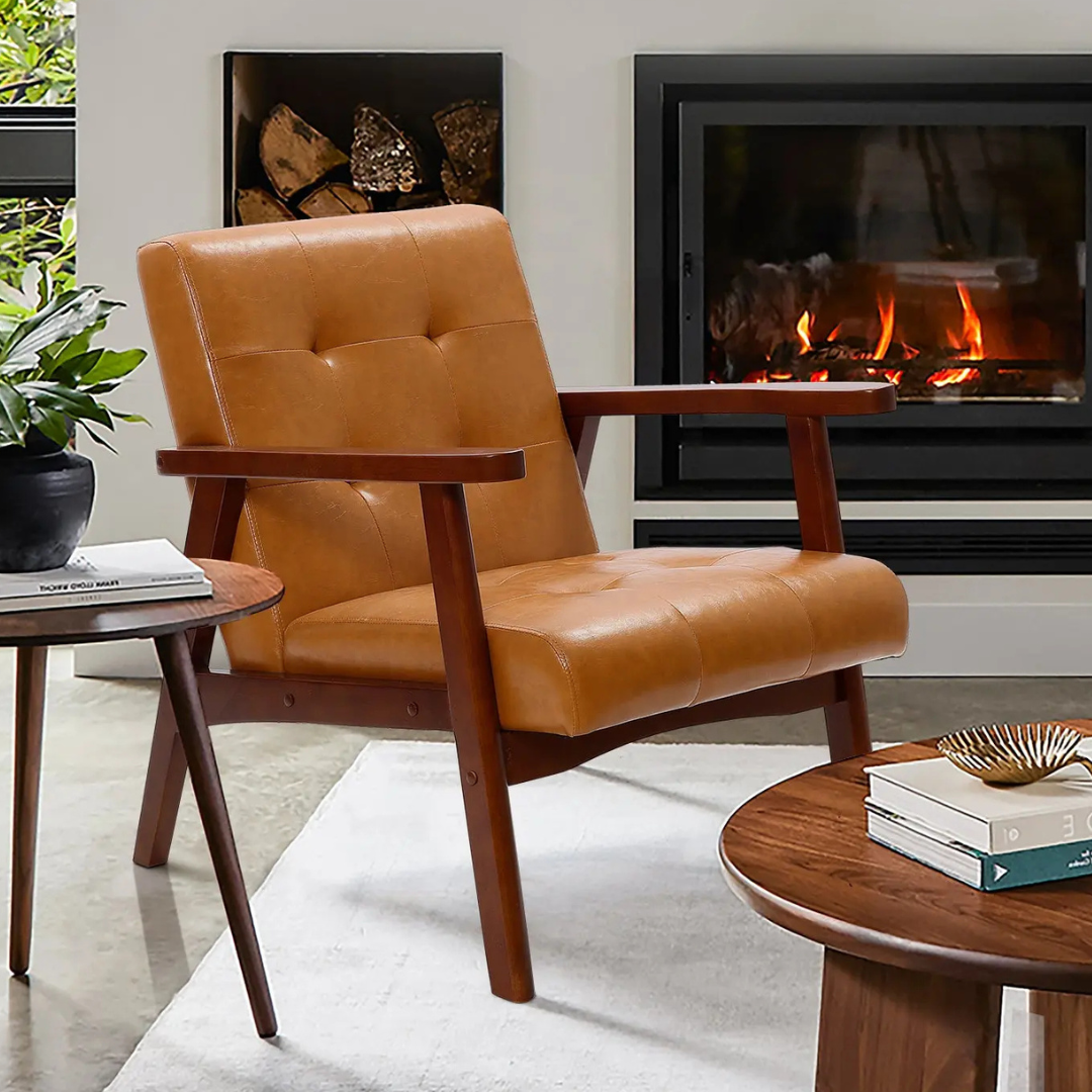 The LuxenHome Josef Accent Chair features a mid-century modern design with a tufted, caramel-colored faux leather seat and backrest. It has wooden arms and legs, set in a warm, cozy living space near a fireplace.