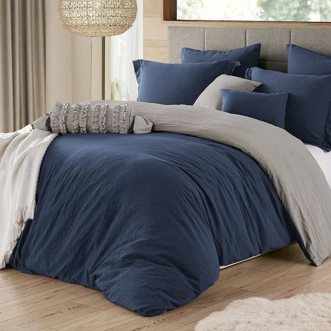 A cozy bedroom scene features a bed with a navy-blue duvet cover, a light gray blanket, multiple matching pillows, and a gray ruffled bolster pillow, all set in a warmly lit room with wooden flooring and a woven hanging light fixture.