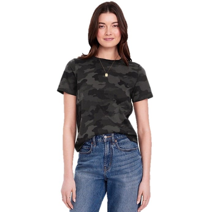 A woman is wearing a dark camo-print t-shirt paired with high-waisted blue jeans. She has a pendant necklace on and is smiling.