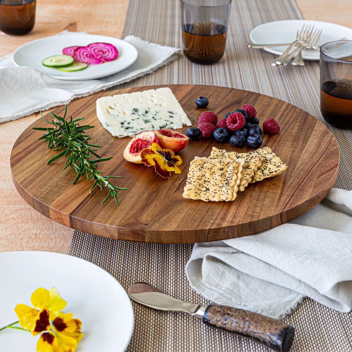 A round wooden serving board with cheese, crackers, rosemary, blueberries, raspberries, blood orange slices, and edible flowers, set on a table with plates and glasses.