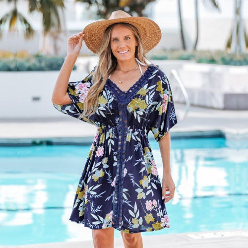 A woman stands by a pool wearing a floral-patterned navy dress with a lace detail and a wide-brimmed straw hat.