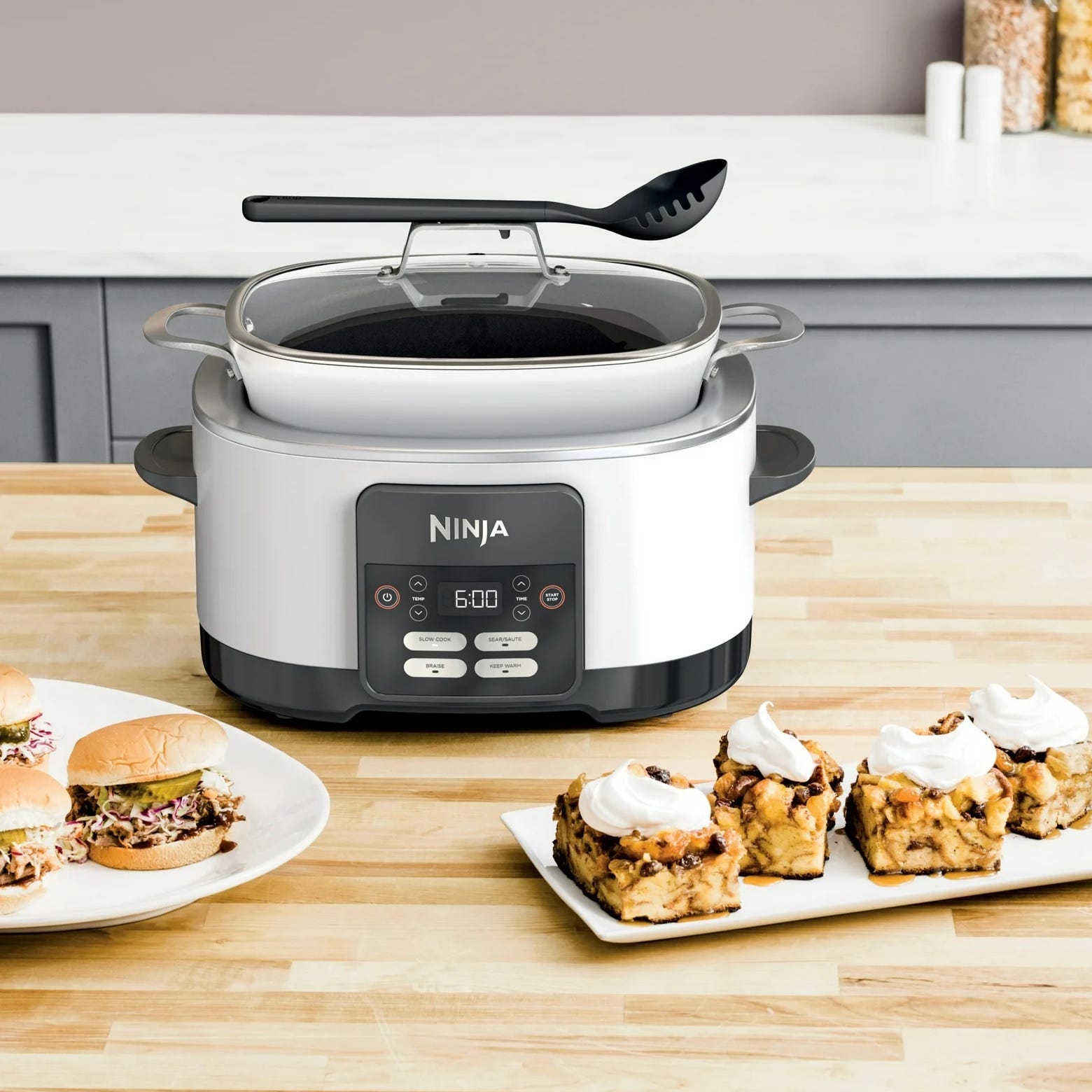 A Ninja multi-cooker on a kitchen counter surrounded by plates of sandwiches and cinnamon rolls.