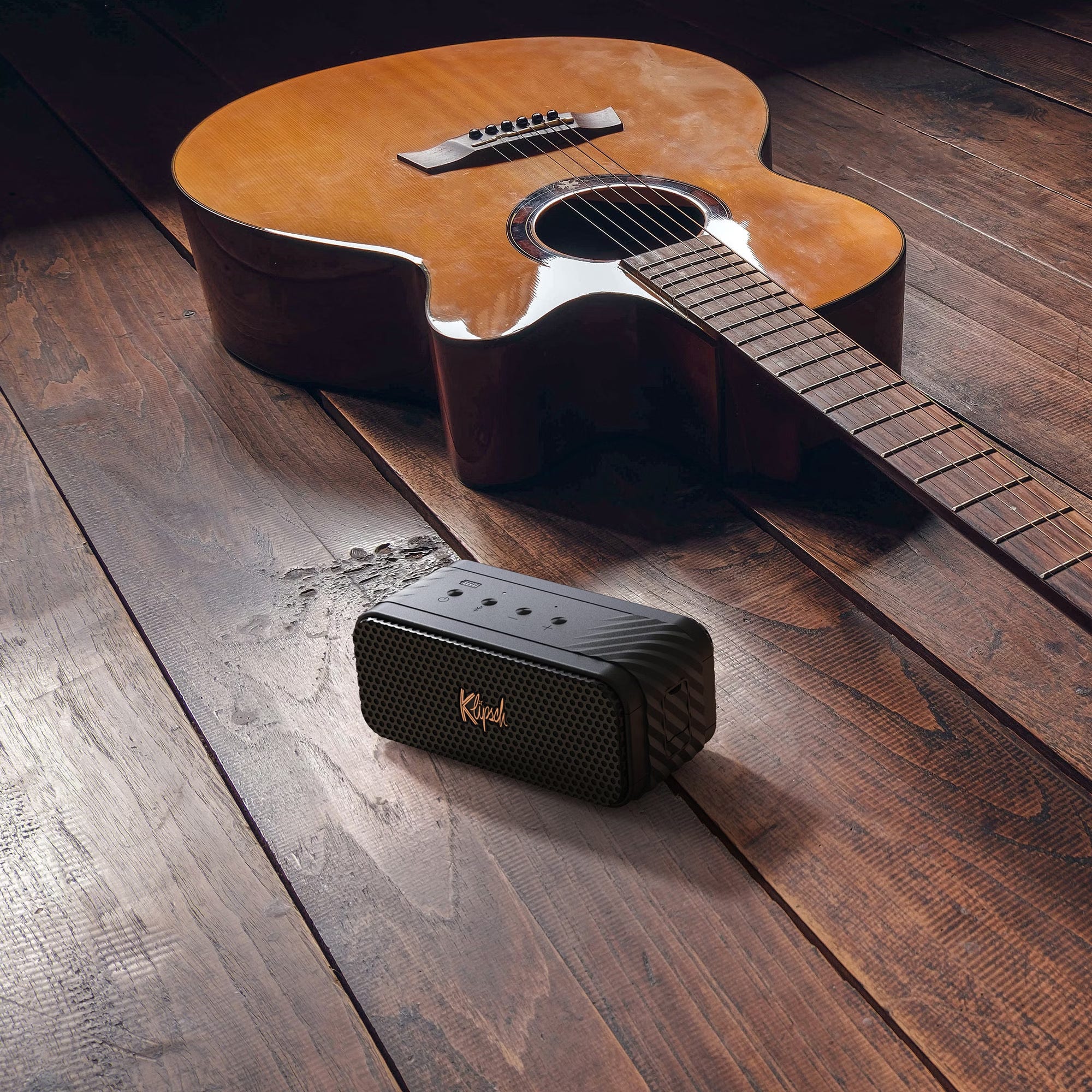 An acoustic guitar next to a portable speaker on a wooden floor.