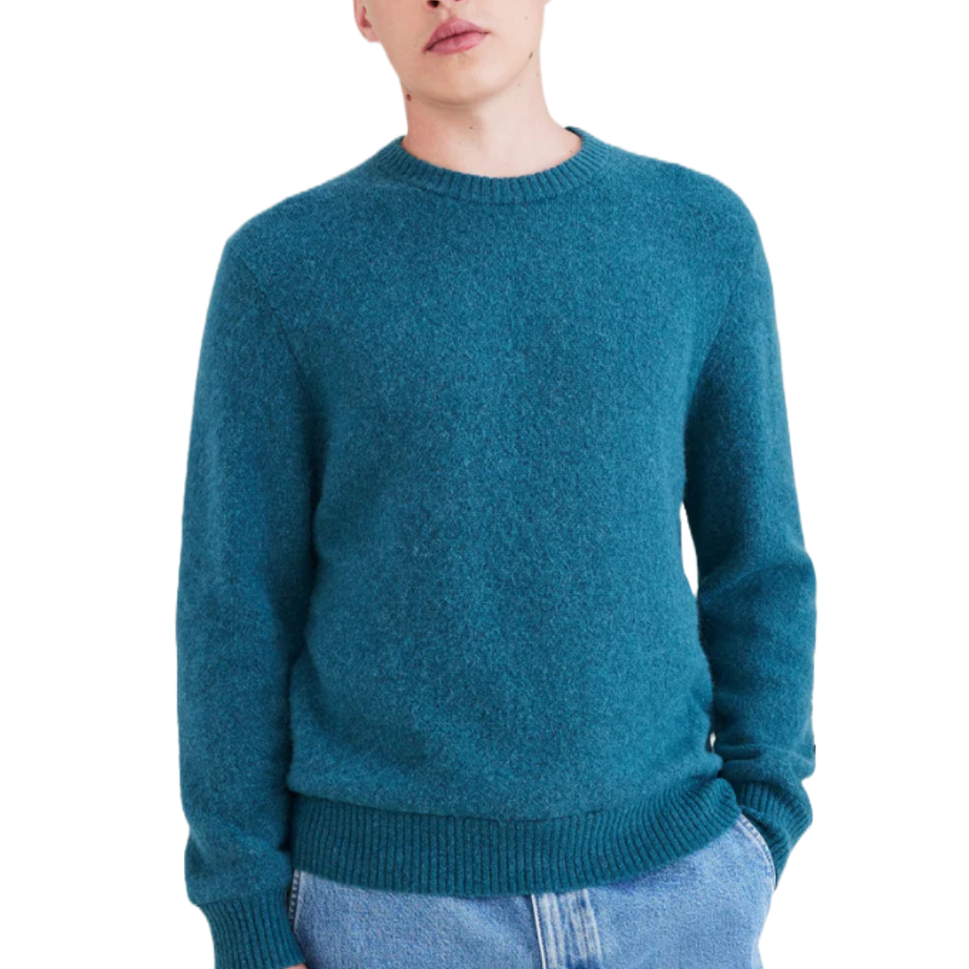 Male model wearing a teal crewneck sweater paired with light blue jeans, hands in pockets.