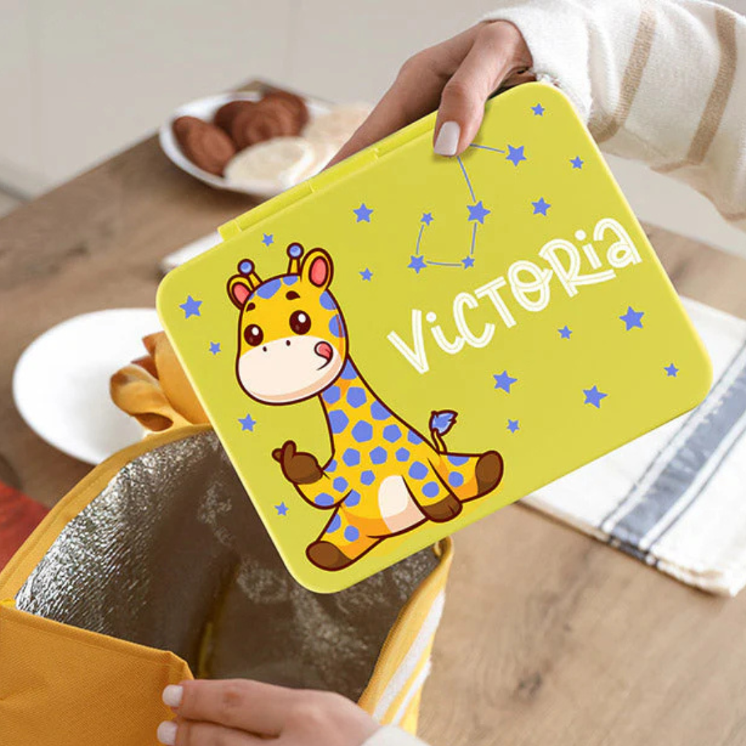 A yellow lunchbox features a cute cartoon giraffe with blue spots and the name \“Victoria\“ in white, surrounded by blue stars.