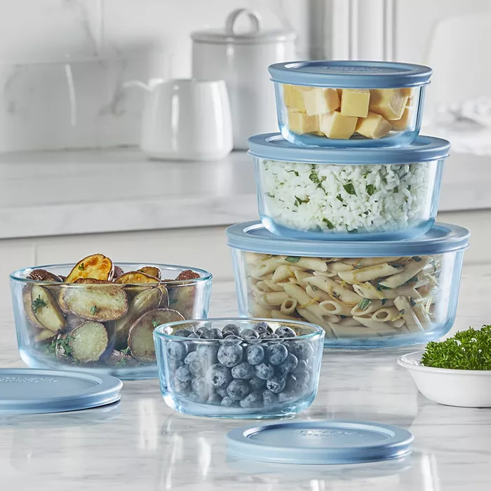 Glass food storage containers with blue lids, containing pasta, rice, cheese, roasted potatoes, and blueberries.