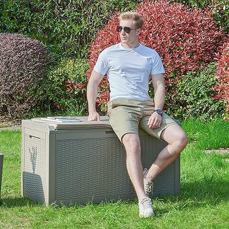 A man in a white T-shirt and shorts is seated on a gray outdoor storage bench in a garden.