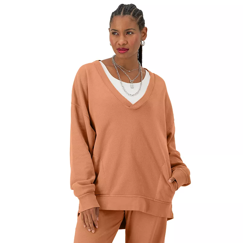 A woman is wearing a camel-colored V-neck sweatshirt and matching sweatpants with layered necklaces.