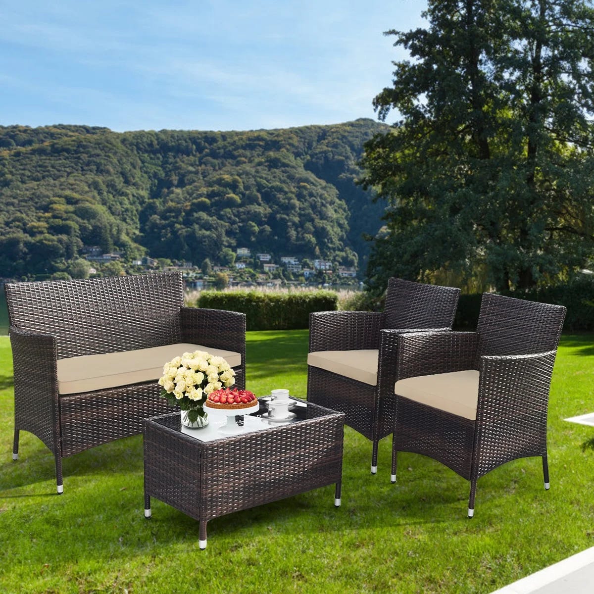 Outdoor wicker patio furniture set with a loveseat, two chairs, and a coffee table. Cushions are cream-colored, and the table holds a vase of flowers and a tray with teacups and fruit. The set is placed on a lush green lawn with a scenic mountain view.