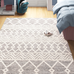 A patterned area rug with a geometric design in shades of beige and dark gray, positioned in a bedroom setting with visible furniture and a pair of shoes on top.