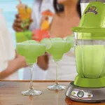 A green Margaritaville drink maker with a blending jar on top is shown next to two margarita glasses filled with a slushy green beverage, garnished with lime slices.