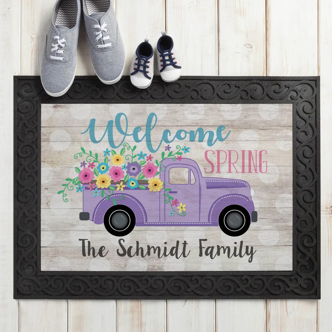 A welcome doormat displays the message \“Welcome Spring,\“ featuring a purple truck filled with colorful flowers, alongside the text \“The Schmidt Family.\“ It's placed on a wooden floor with a decorative black border, with adult and child shoes nearby.
