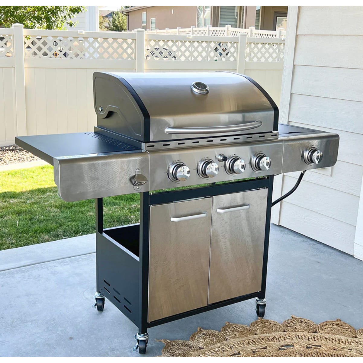Stainless steel gas grill with five burners, side shelves, and a lower cabinet on wheels, set outdoors on a patio.