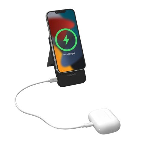 A charging stand holds a smartphone with a visible charging icon, while a cable connects the stand to a pair of wireless earbuds, indicating simultaneous charging.