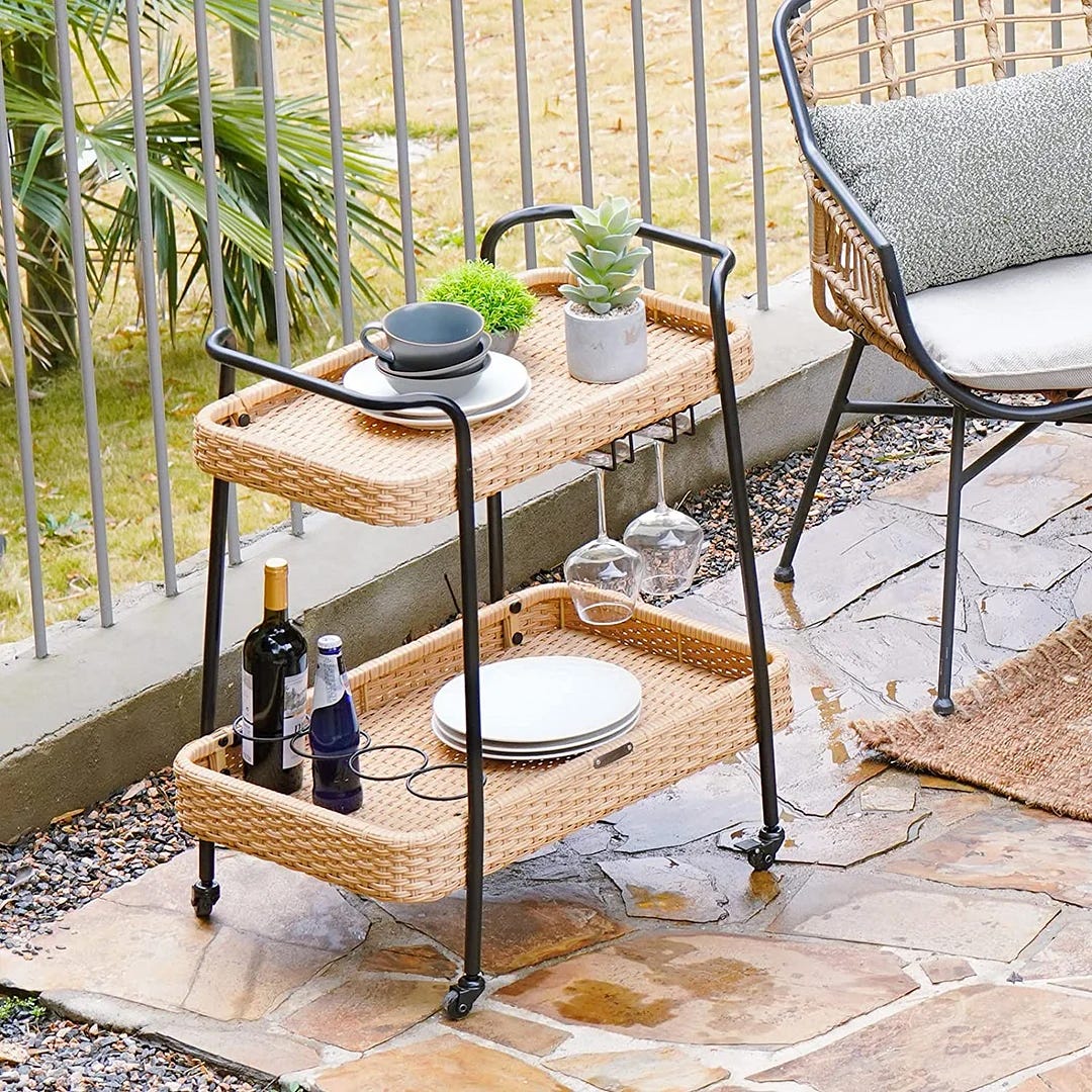 A two-tiered rattan serving cart with dishes, plants, and a wine bottle displayed on it, situated on a patio beside an outdoor chair.