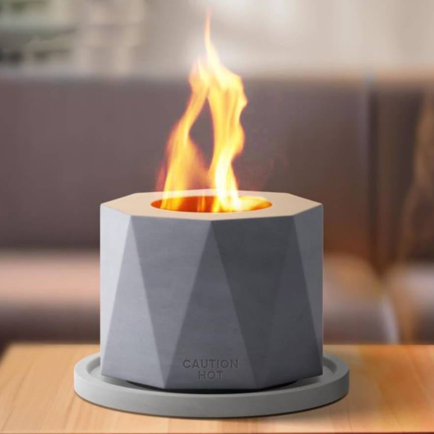 A tabletop portable fire pit with visible flames, set against a blurred indoor background.
