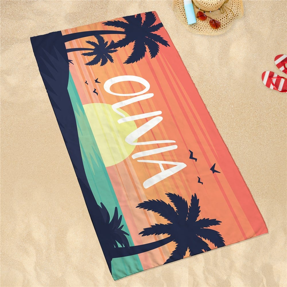Personalized quick-dry beach towel with \“Olivia\“ in white text on a sunset background featuring silhouettes of palm trees and birds. It's displayed on sand with a hat, sunscreen, sunglasses, and flip-flops nearby.