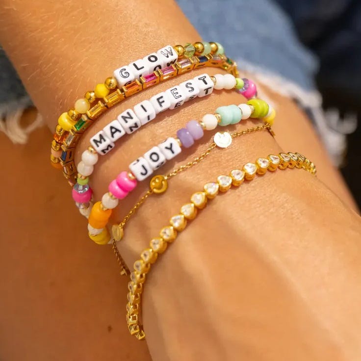 A wrist adorned with colorful beaded bracelets, some featuring words like \“GLOW,\“ \“MANIFEST,\“ and \“JOY.\“