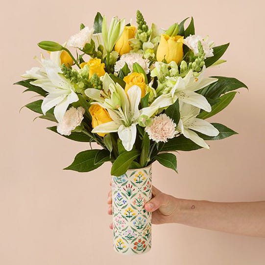 A bouquet of yellow roses, white lilies, and other mixed flowers held in a patterned vase.