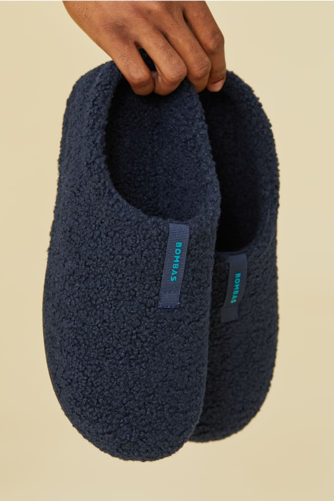 A pair of dark-colored, fuzzy Bombas Sunday Slippers with a tag on the side are being held up.
