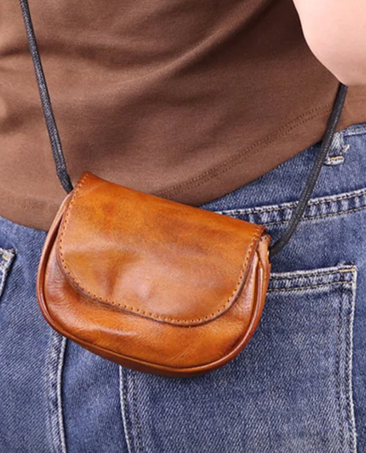 A small, brown leather crossbody bag with a flap closure, worn over a brown shirt and blue jeans.