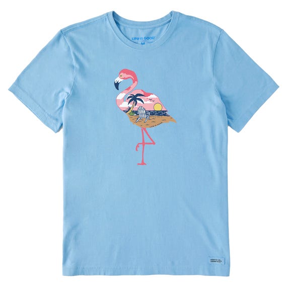 Light blue t-shirt featuring a graphic of a pink flamingo standing on one leg, with a tropical scene inside its body, including palm trees and a sunset. \“Life is Good\“ is printed at the collar.