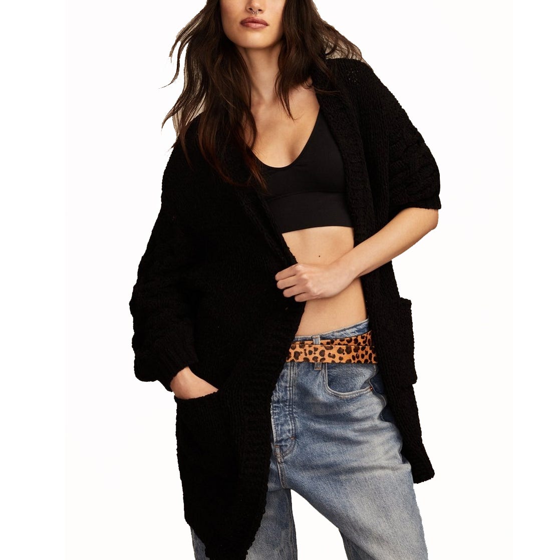 A woman wears a black knitted cardigan over a black crop top, paired with loose blue jeans and a leopard print belt.