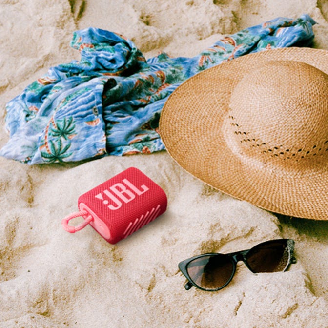 A red portable speaker, straw sun hat, sunglasses, and a blue patterned shirt on sand.