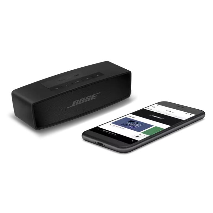 A Bose portable speaker next to a smartphone displaying music playback controls.