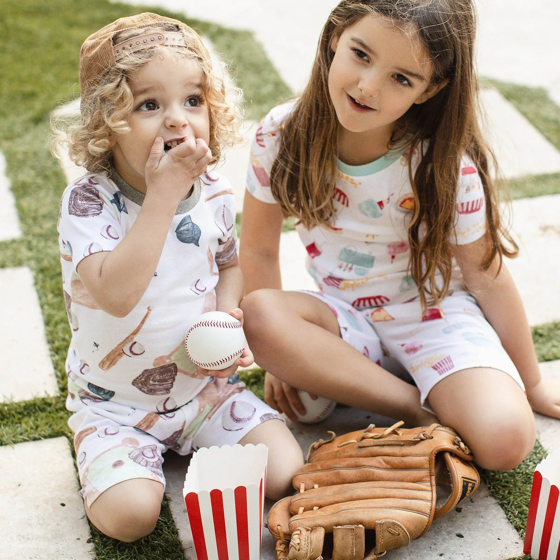 Two children wearing baseball-themed pajamas, one holding a baseball, sit outdoors next to a popcorn box and baseball glove on grass.