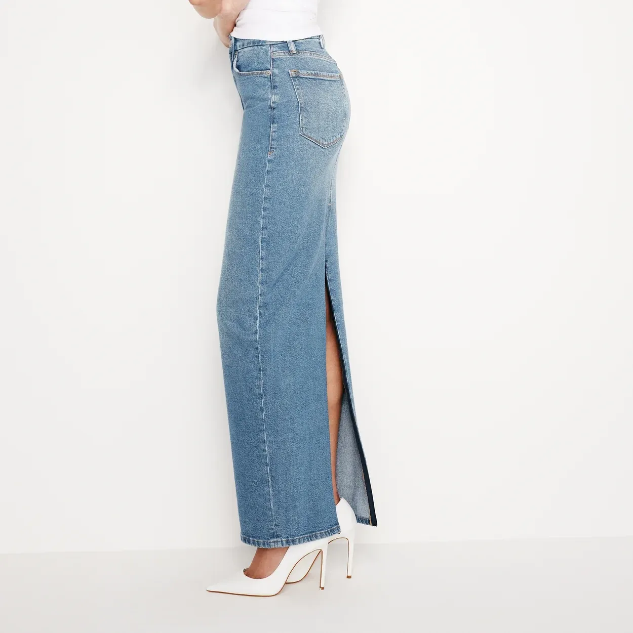 A denim maxi skirt with a high waist and front slit that goes up the thigh, paired with white high-heeled shoes.