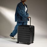 A person is walking with a black Antler Luggage Icon Stripe Spinner, which features a hard shell design, horizontal stripes, and four wheels.