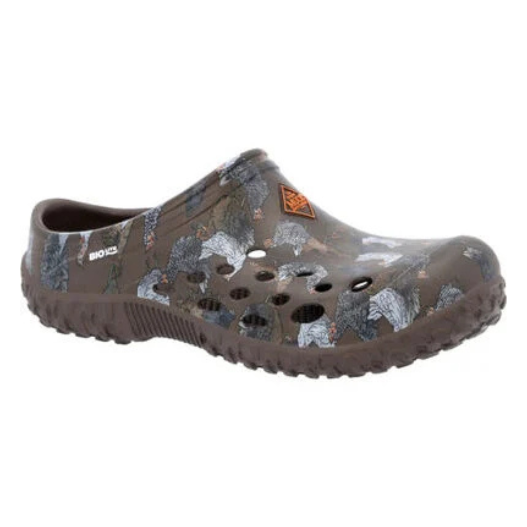 Muck Boot Eva Clogs are brown slip-on footwear with a camouflage pattern featuring trees and animals. They have ventilation holes and rugged soles for outdoor use.