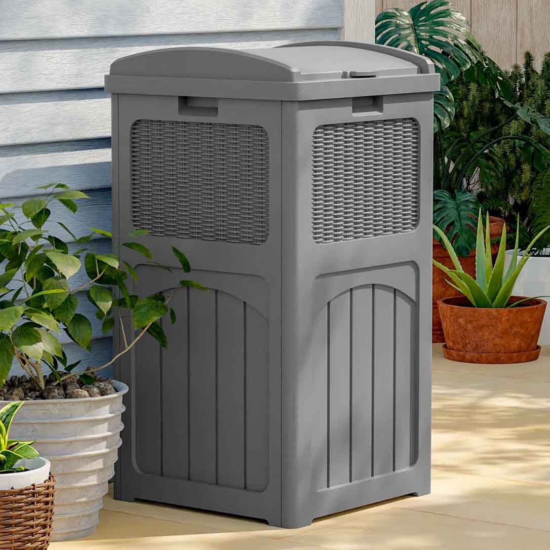 A gray outdoor trash can with a wicker design detail stands on a patio, surrounded by various potted plants.
