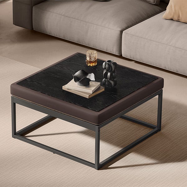A square coffee table with a black wood top and brown leather edges, on a metal frame, with a glass, a book, and decorative object on it.
