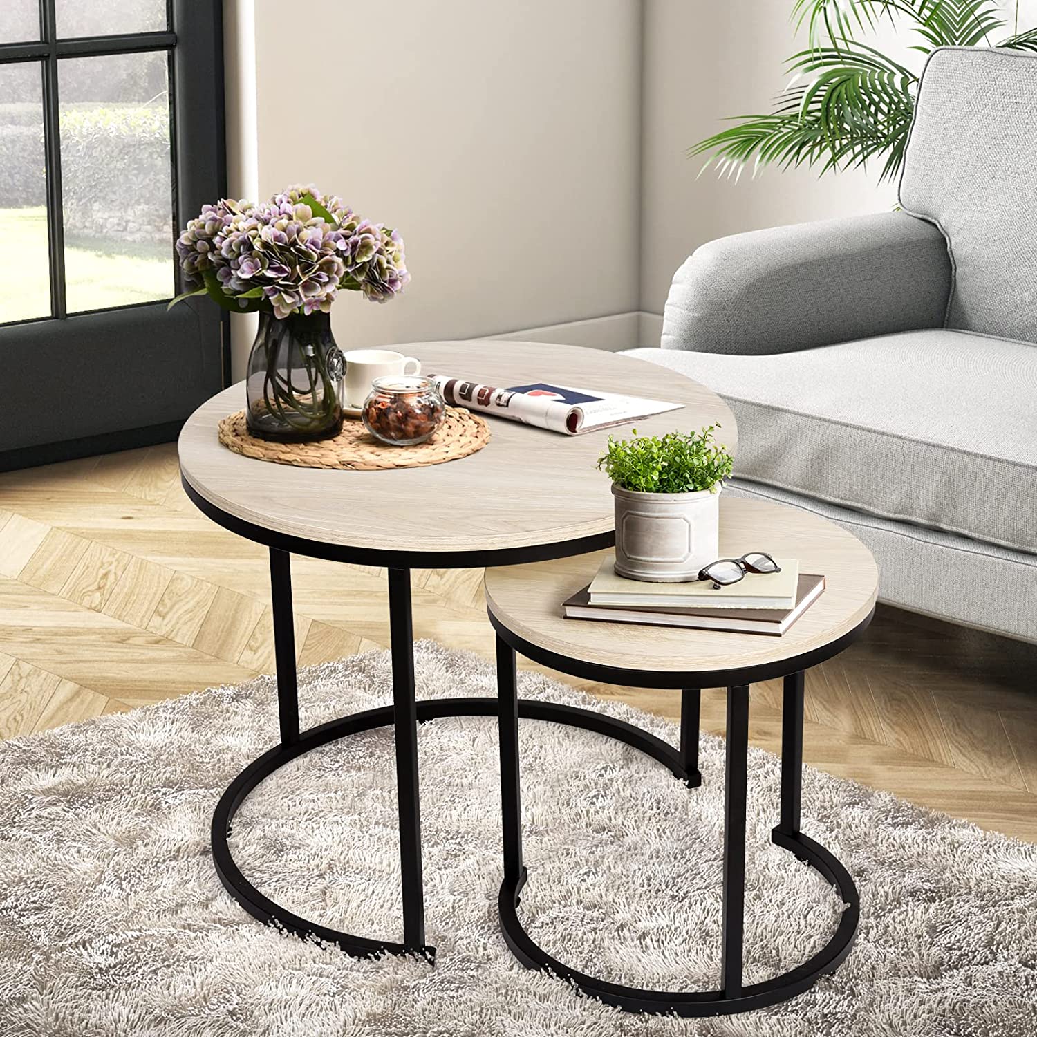 Two round nesting tables with black metal frames and light wood tabletops, one larger than the other, arranged in a living room setting.