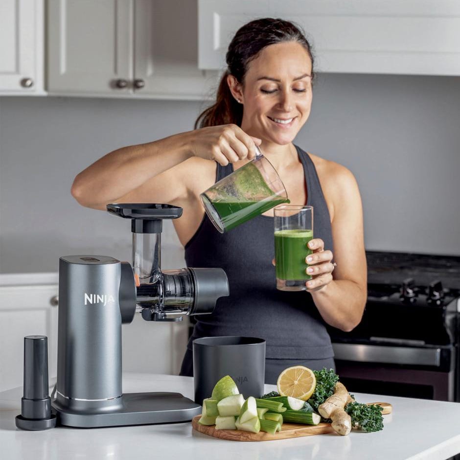 A woman pours green juice into a glass beside a Ninja brand juicer and fresh produce like kale, lemon, and cucumber.