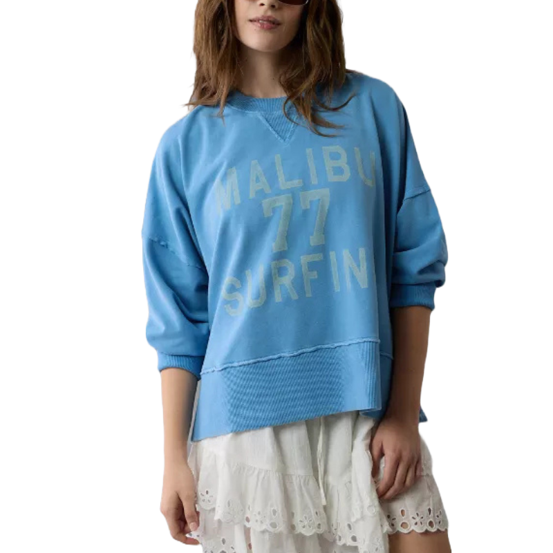 Blue oversized sweatshirt featuring \“MALIBU 77 SURFIN'\“ text, with ribbed cuffs and hem, worn over a white eyelet lace mini skirt.
