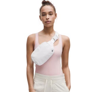 A woman with a top bun hairstyle wears a light pink sleeveless bodysuit and beige pants, accessorized with a white crossbody belt bag.