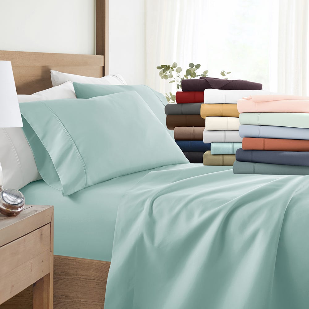 A bed with light teal sheets and pillows is displayed in a bedroom setting, alongside a stack of folded sheets in various colors including red, white, beige, and blue.