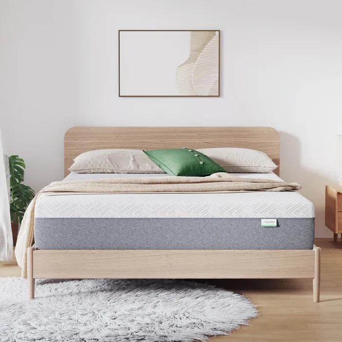 A modern bedroom setup featuring a light wood bed frame, a layered mattress, and neutral bedding with a green decorative pillow. A minimalist painting and a textured gray rug complete the room's aesthetic.