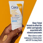 A hand holds a tube of CeraVe Hydrating Mineral Sunscreen SPF 30 with a sheer tint, next to a swatch of the product on the hand.