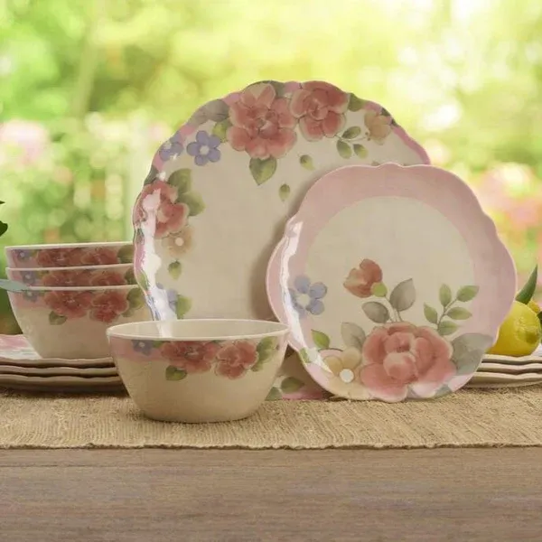 The Pfaltzgraff Tea Rose Dinnerware Set features plates and bowls with scalloped edges, adorned with pink roses and small blue flowers on a beige background. The set has a floral, vintage-inspired design suitable for casual or formal dining.