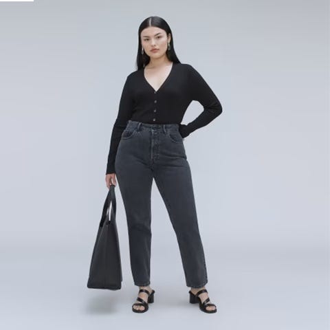 A woman is wearing a black V-neck cardigan, faded black jeans, black strappy sandals, and carrying a large black tote bag.