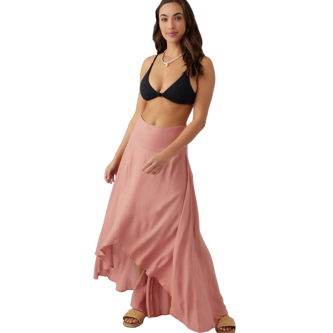 A woman is wearing a black bikini top paired with a pink, flowy asymmetrical maxi skirt and woven flat sandals. She accessorizes with a simple pearl necklace.