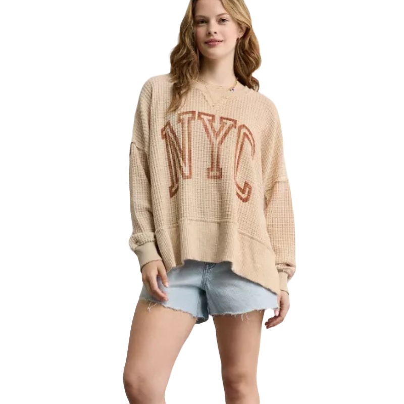 A person wearing a beige oversized sweater with \“NYC\“ printed on it and light denim shorts.