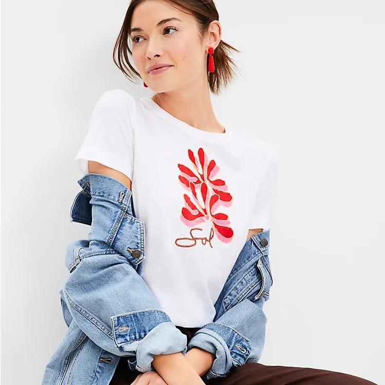 A woman wearing a white T-shirt with a red coral design and cursive text, paired with a denim jacket and red earrings.